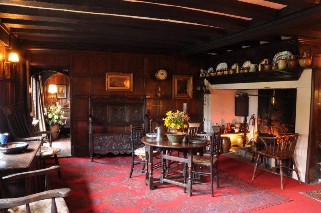The Monks Parlour with beams, wood paneled walls and a large inglenook fireplace, red carpets and a table set with flowers and bowls