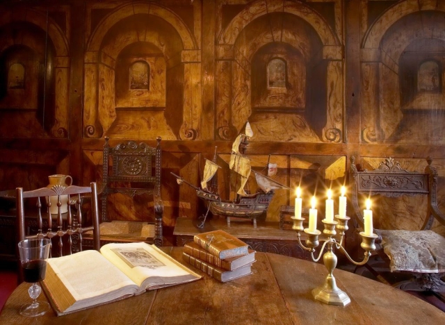 The painted room with trompe l'œil walls depicting wooden recessed areas with small arched windows, the is also a table sith boks and a glass of wine and a lit candleabra and a model ship and ornate carded chairs in te background